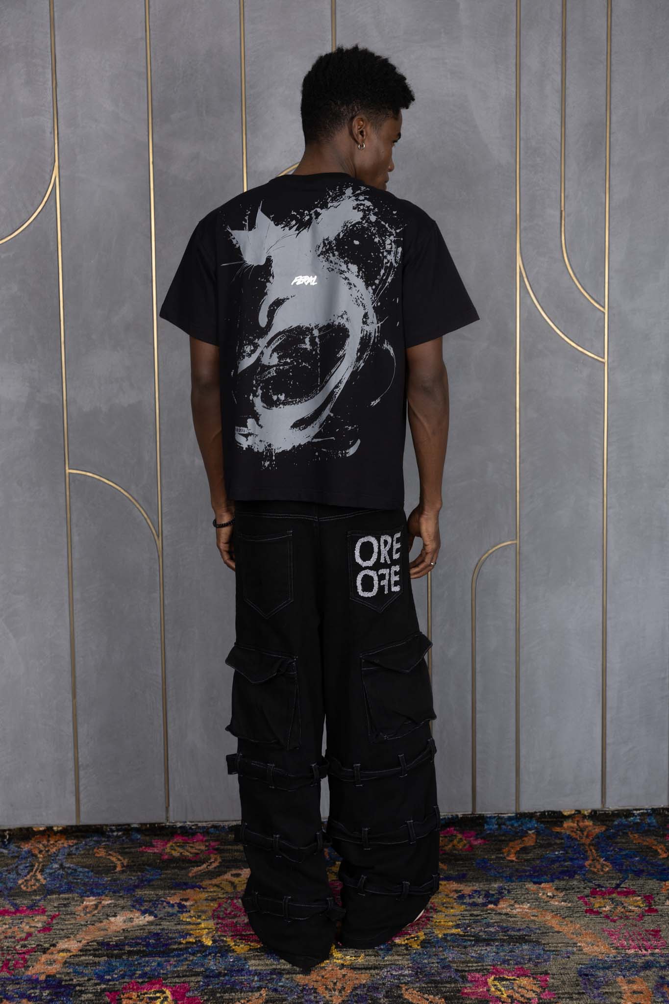 Person wearing ORE OFE's blackout t-shirt & denim and standing in front of a grey wall on a textured rug
