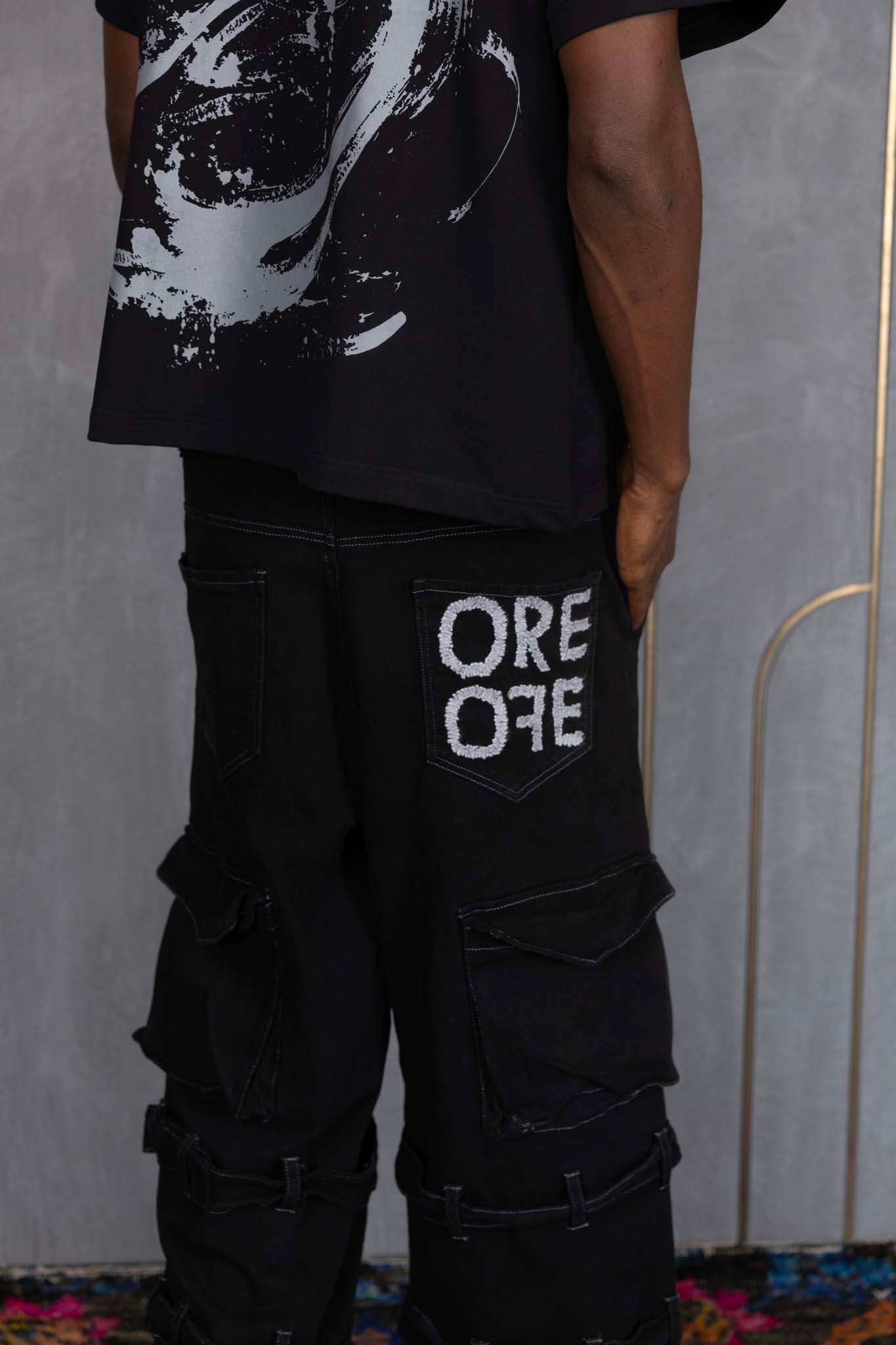 Person wearing ORE OFE's blackout t-shirt & denim and standing in front of a grey wall on a textured rug