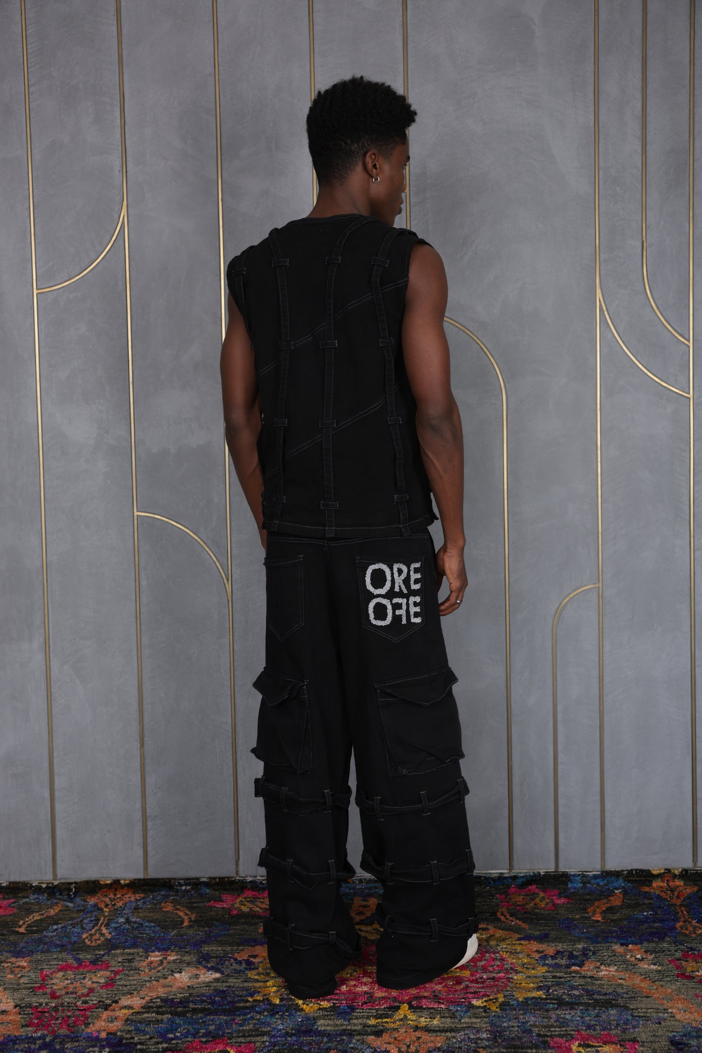 Person wearing ORE OFE's blackout vest & denim and standing in front of a grey wall on a textured rug
