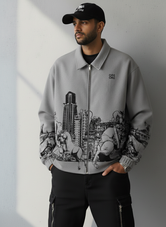 Man wearing a gray jacket with a cityscape design, black cap, and black pants against a light background.