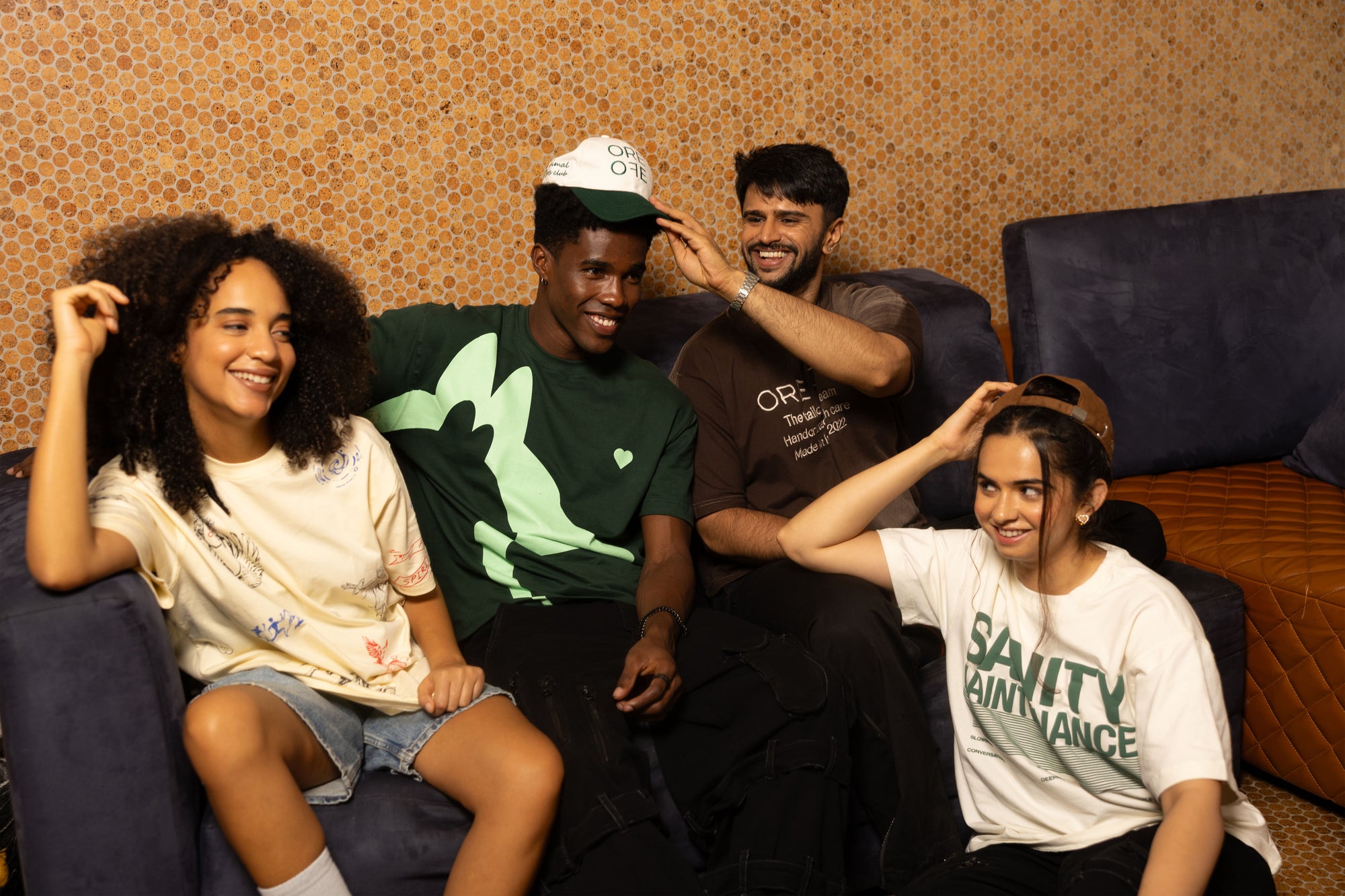 Four friends sitting together on a couch with a patterned wall in the background wearing ORE OFE oversized t-shirts and caps
