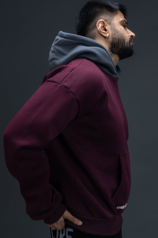 Person wearing a maroon contrast hoodie by ORE OFE with chest & back puff printed design standaing against a dark grey background