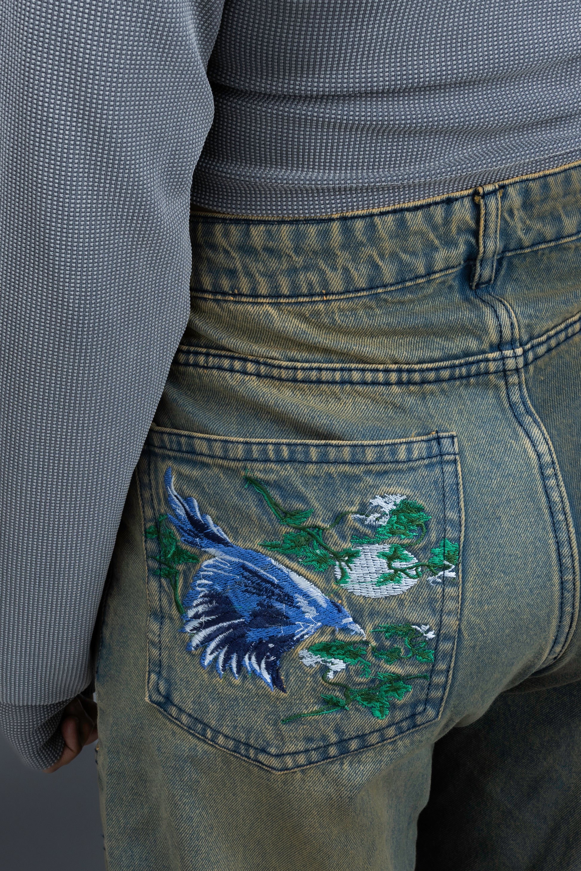 Person wearing ORE OFE's Vintage washed denim that has a pheonix embroided on the back pocket standing against a grey background