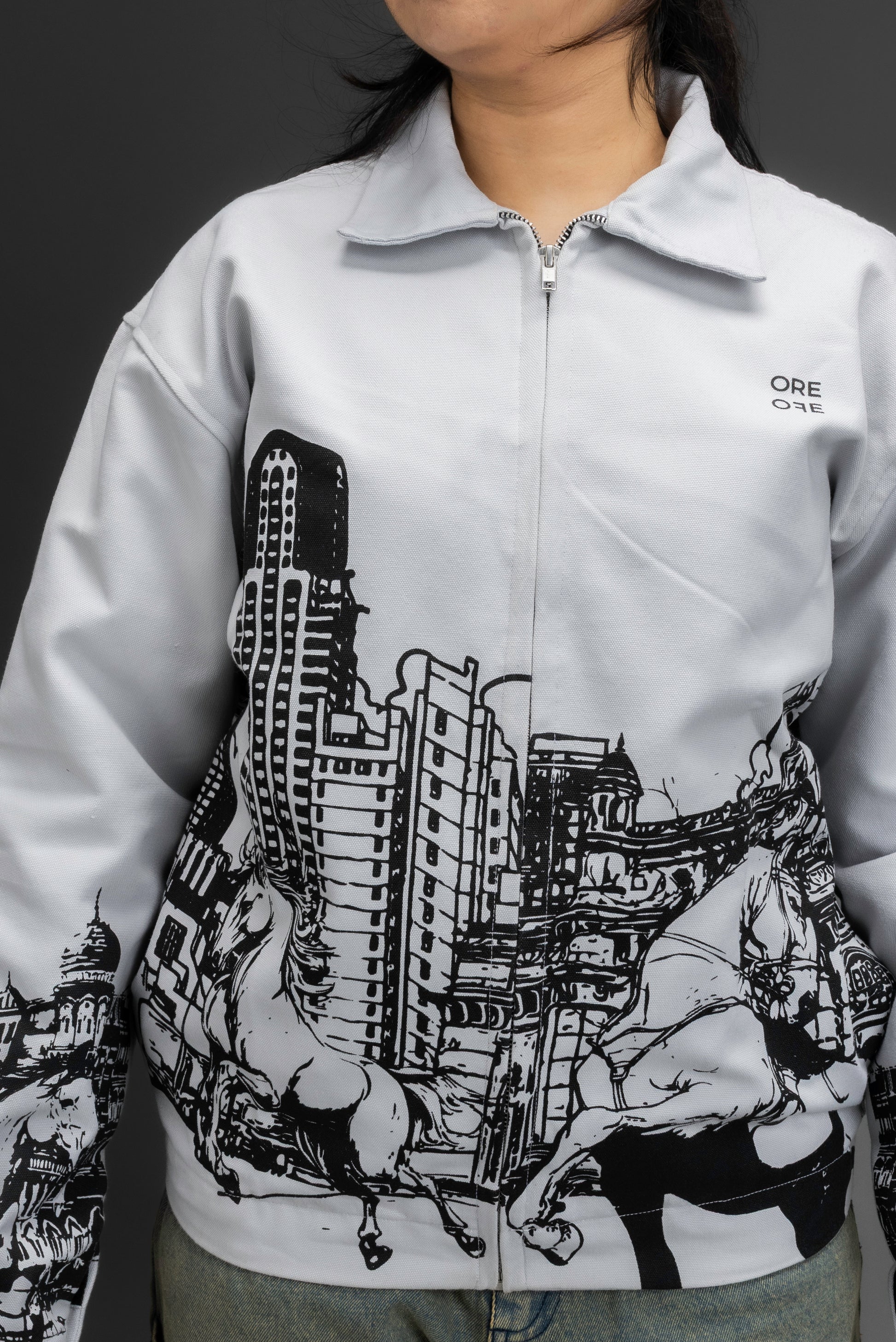 Person wearing a grey jacket with an upper-left chest logo and black cityscape graphic, standing against a dark background