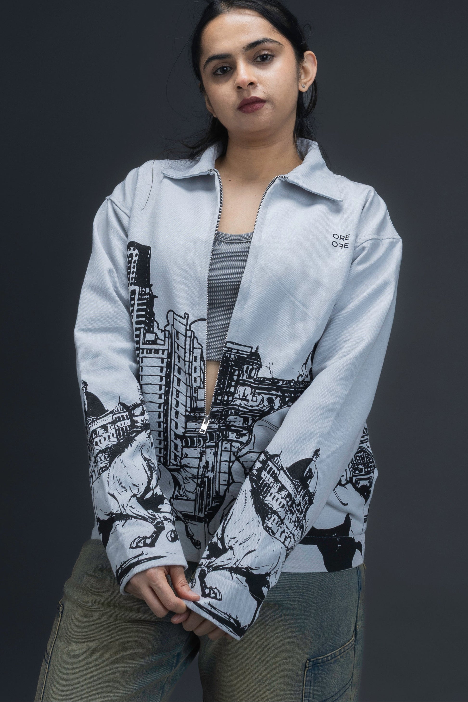 Women wearing a grey jacket with an upper-left chest logo and black cityscape graphic, standing against a dark background