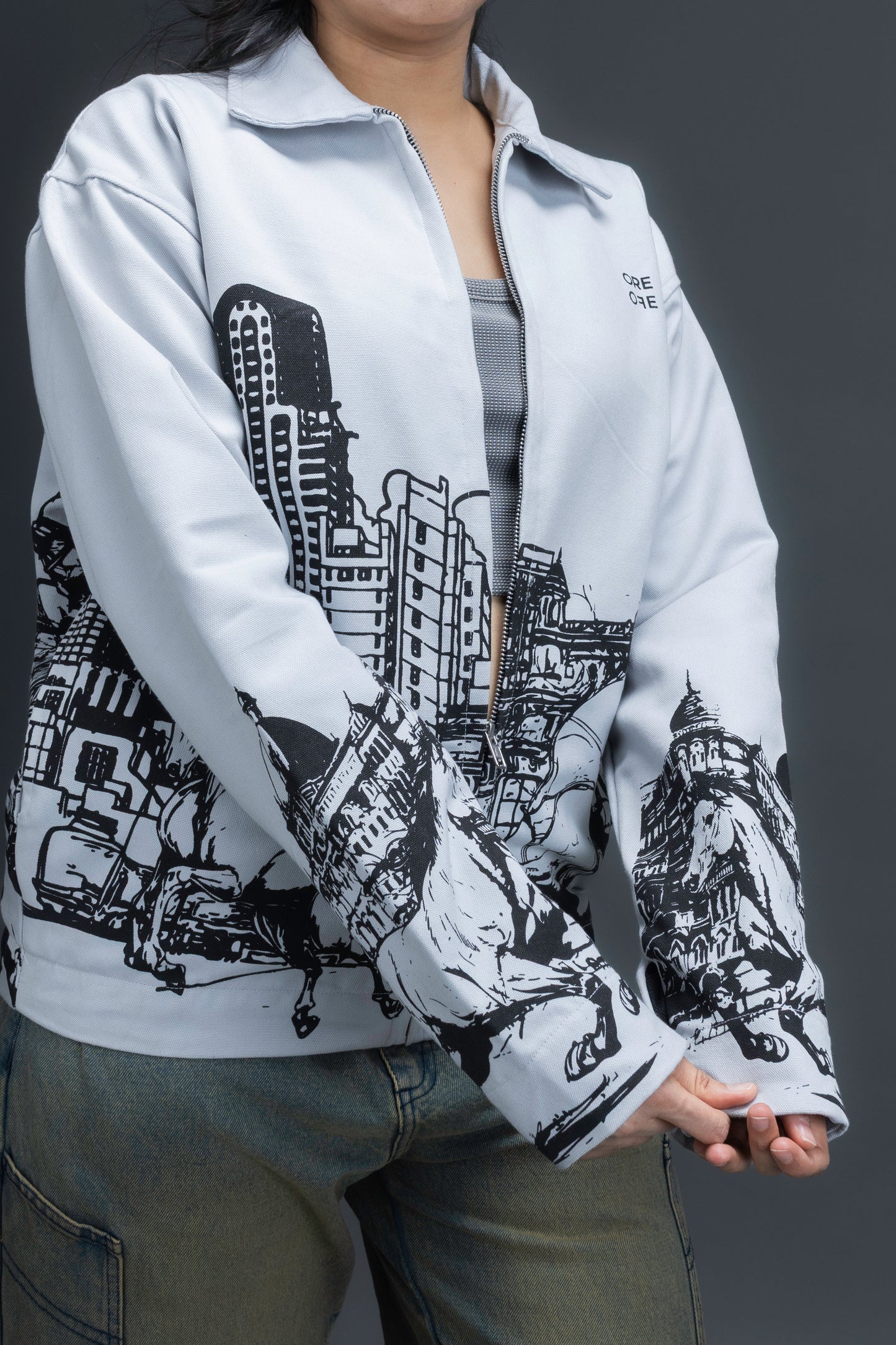 Women wearing a grey jacket with an upper-left chest logo and black cityscape graphic, standing against a dark background