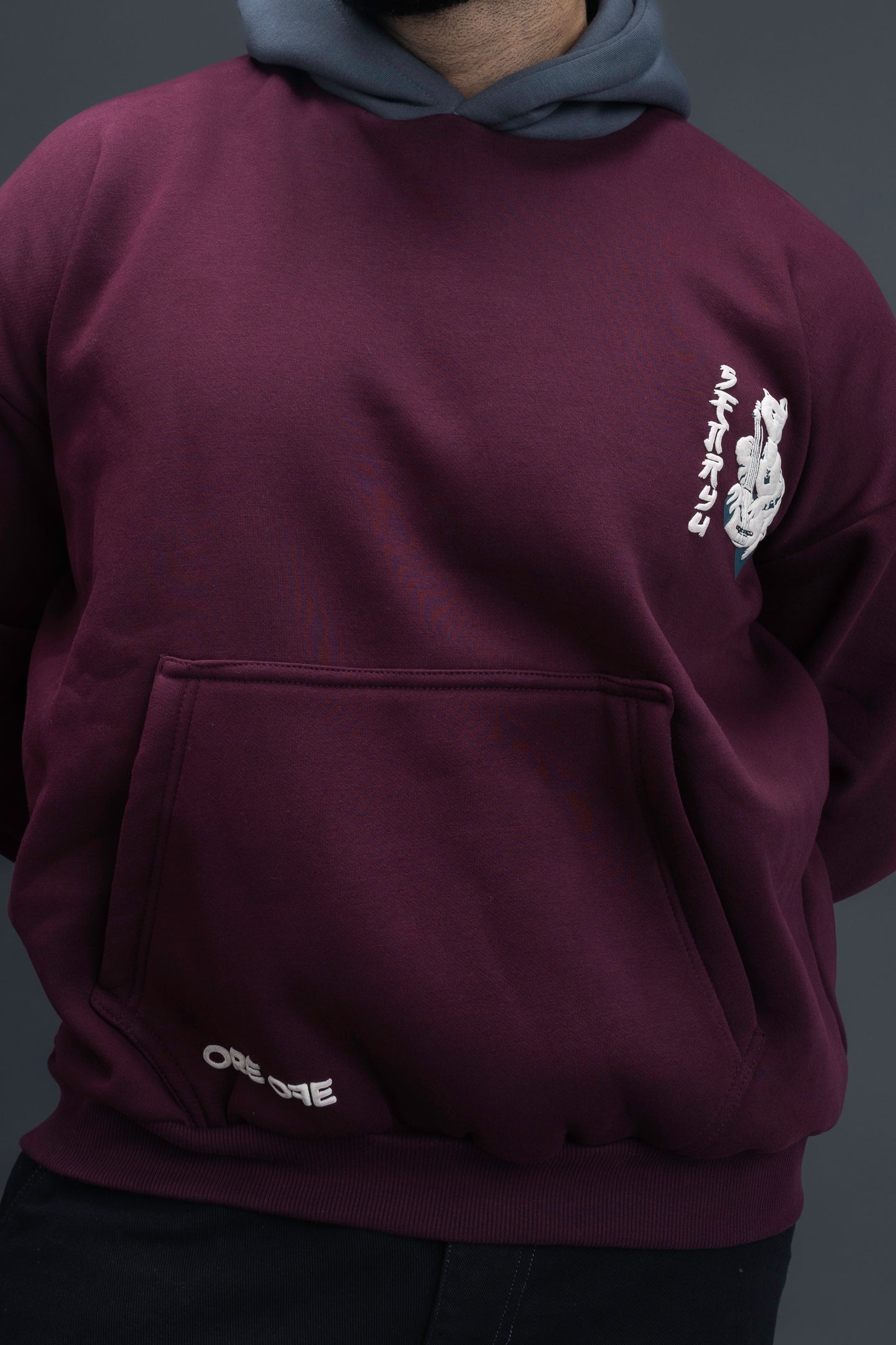 Person wearing a maroon contrast hoodie by ORE OFE with chest & back puff printed design  standaing against  a dark grey background