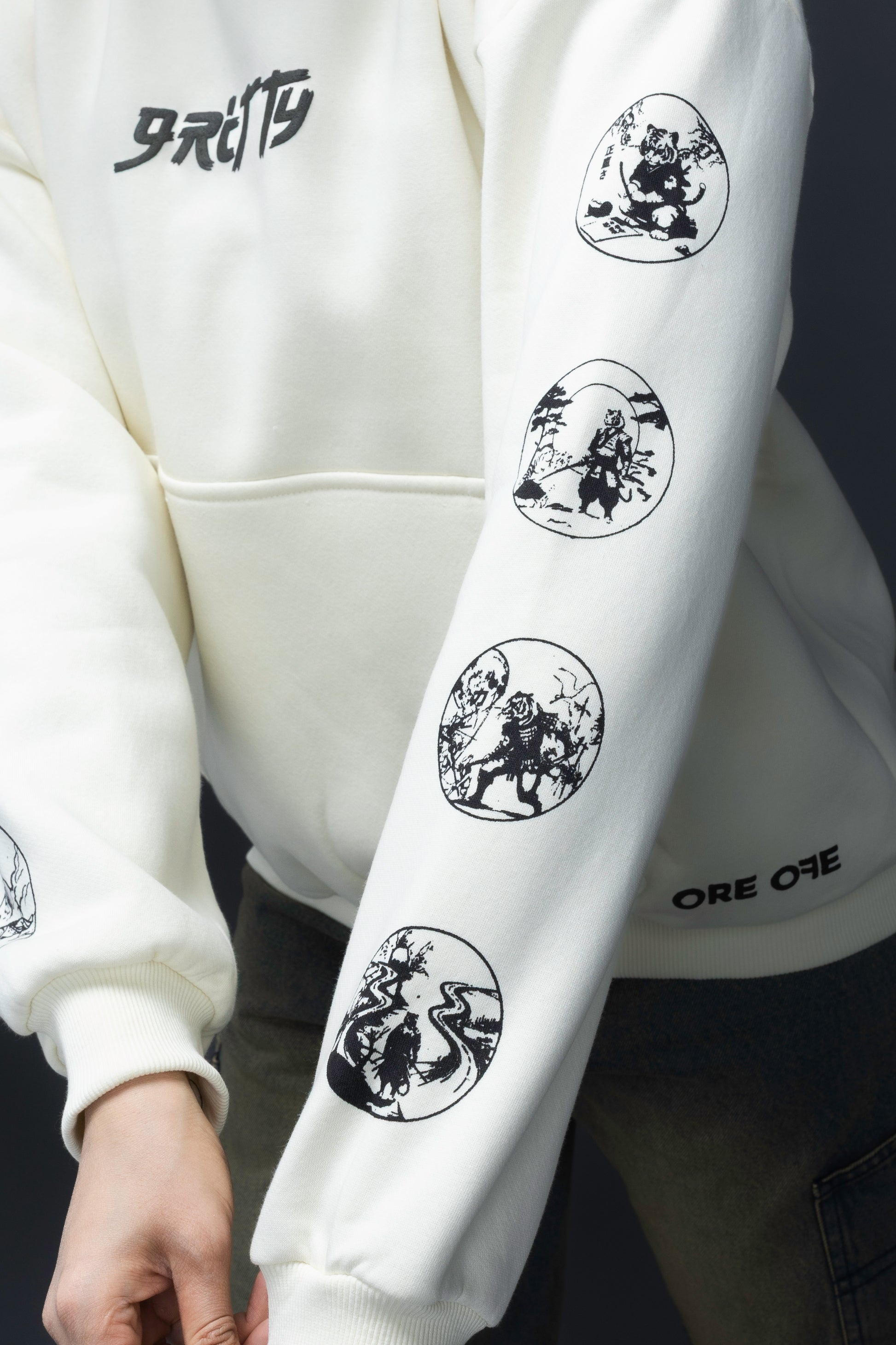 Person wearing a off-white contrast hoodie by ORE OFE with text on the chest that reads GRITTY & design on both sleeves, standing against a black background 