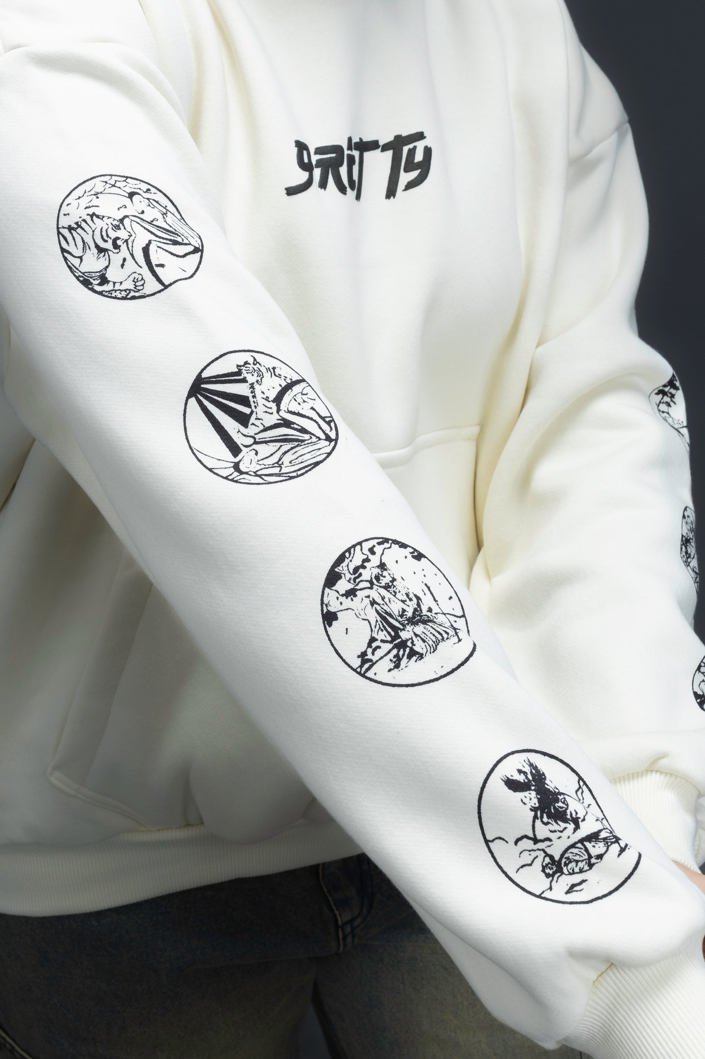 Person wearing a off-white contrast hoodie by ORE OFE with text on the chest that reads GRITTY & design on both sleeves, standing against a black background 