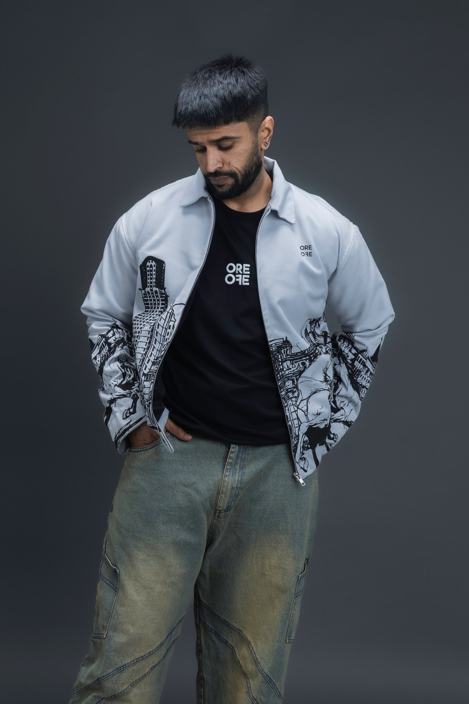 Person wearing ORE OFE's light grey jacket that has the Bombay skyline with horses printed on it standing against a dark grey background