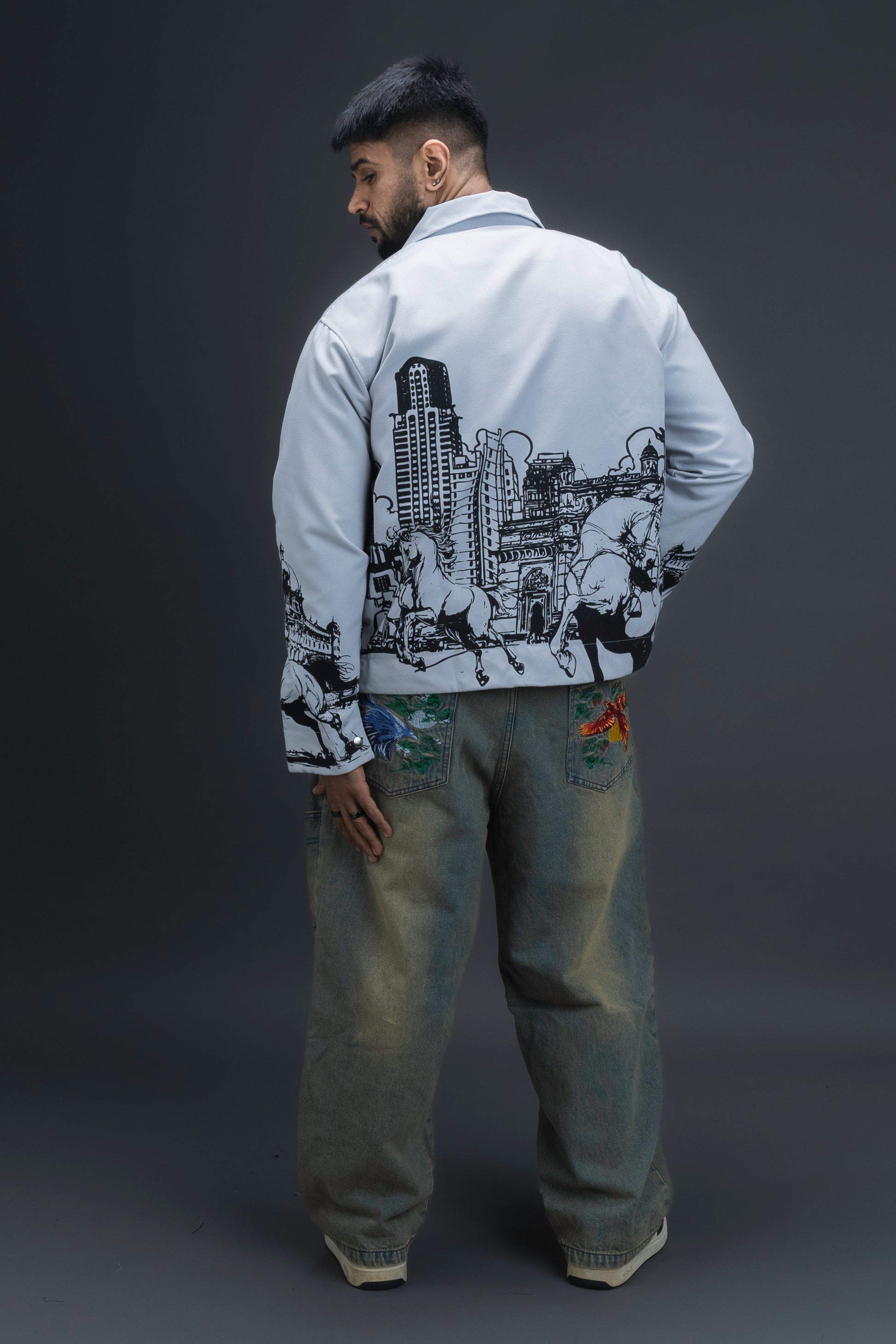 Person wearing ORE OFE's light grey jacket that has the Bombay skyline with horses printed on it standing against a dark grey background