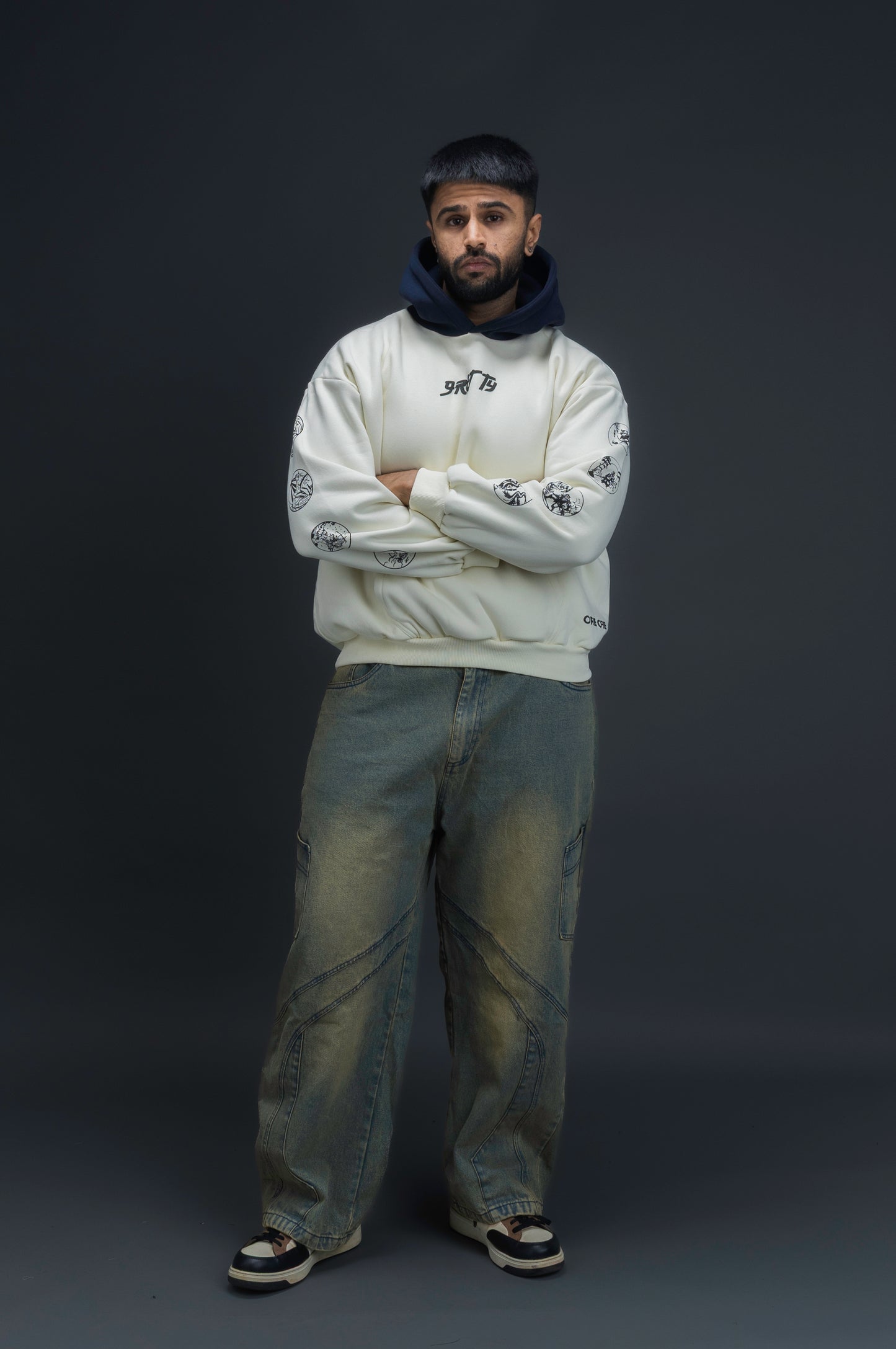 Person wearing a off-white contrast hoodie by ORE OFE with text on the chest that reads GRITTY & design on both sleeves, standing against a black background 