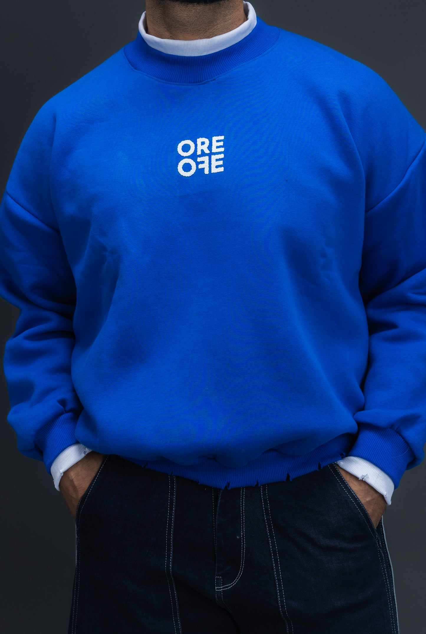 Man wearing ORE OFE's blue contrast sweatshirt with the logo on the centre of the chest standing against a grey background