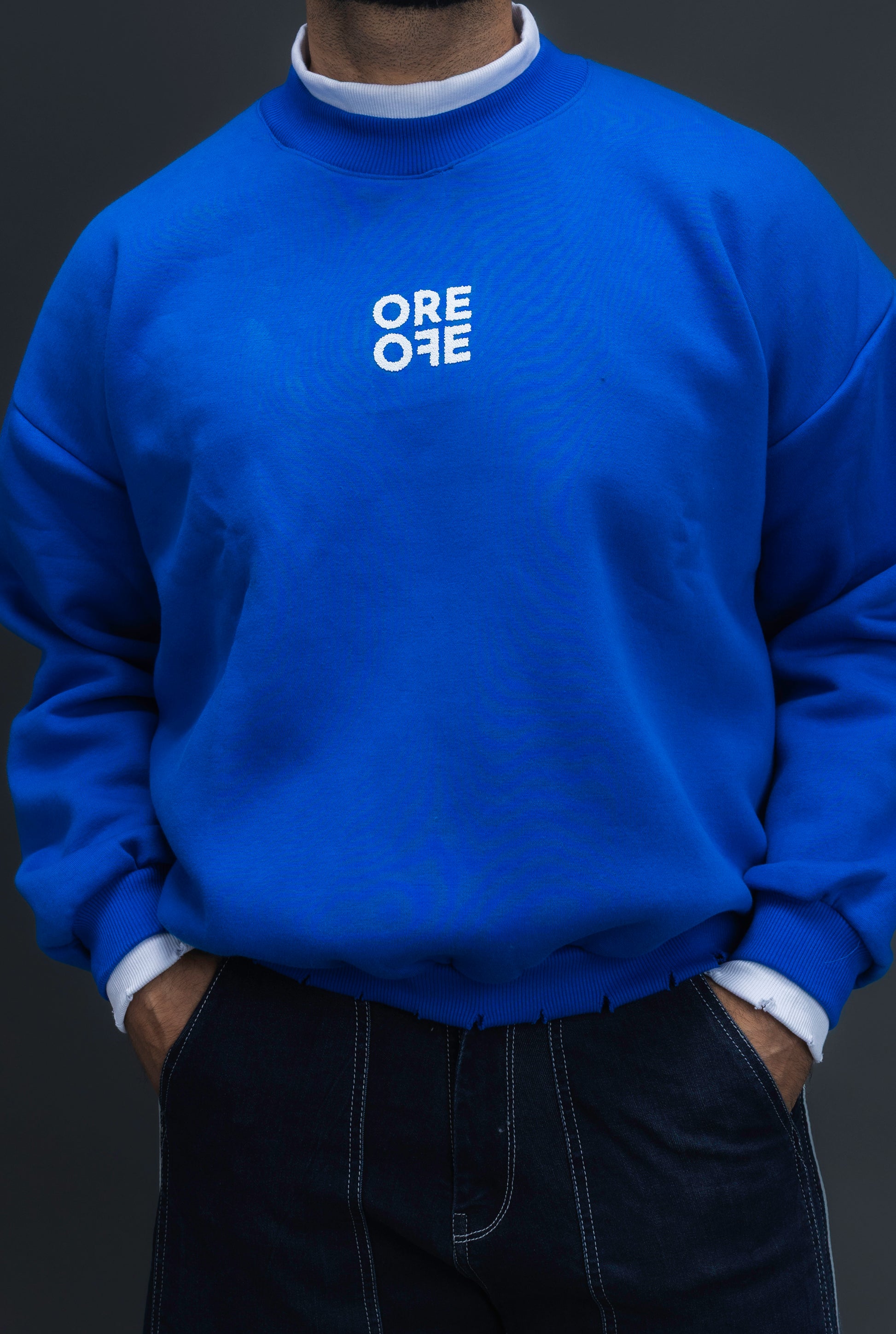 Man wearing ORE OFE's blue contrast sweatshirt with the logo on the centre of the chest standing against a grey background