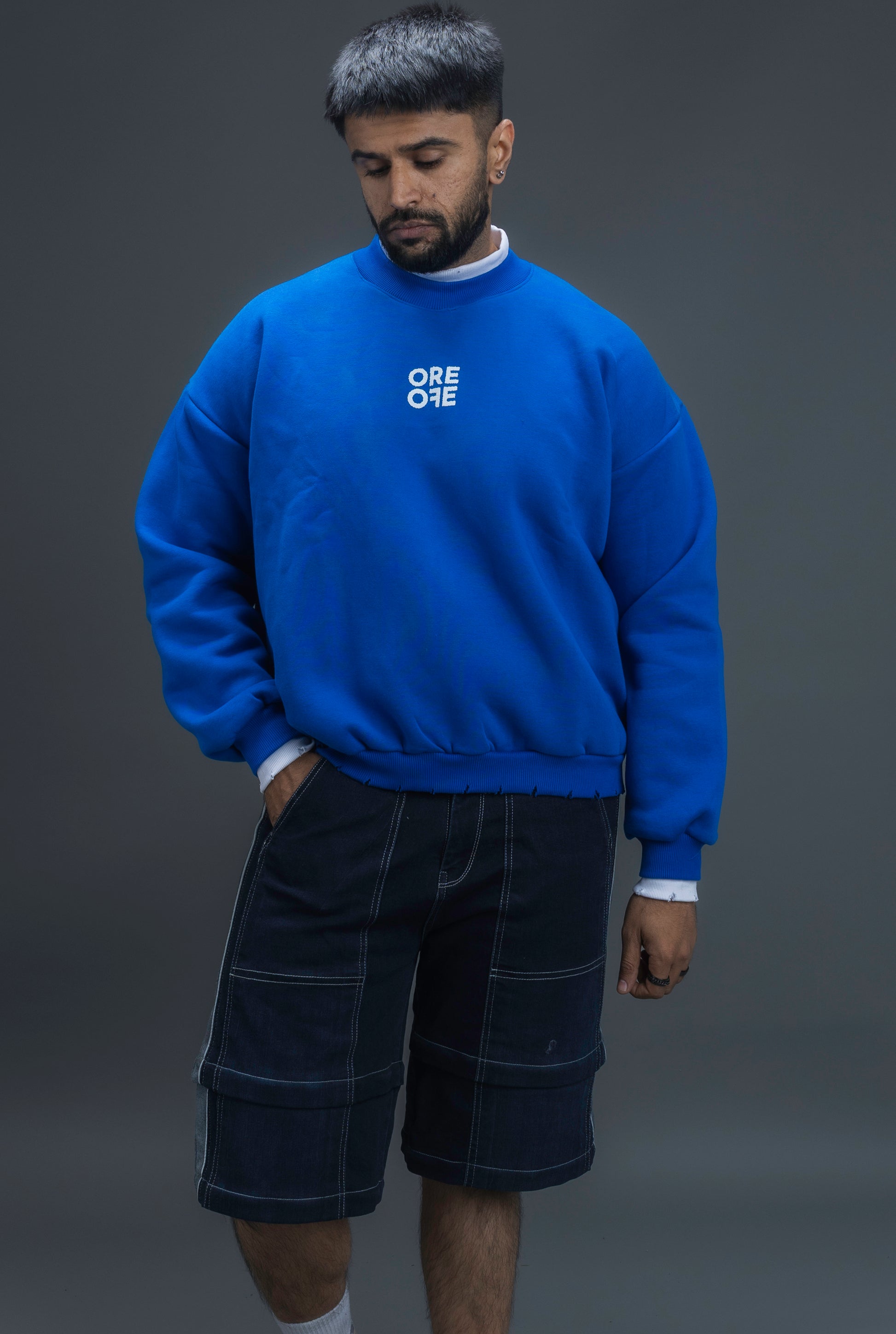 Man wearing ORE OFE's blue contrast sweatshirt with the logo on the centre of the chest standing against a grey background