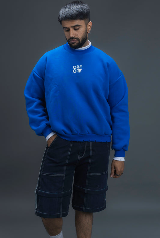 Man wearing ORE OFE's blue contrast sweatshirt with the logo on the centre of the chest standing against a grey background
