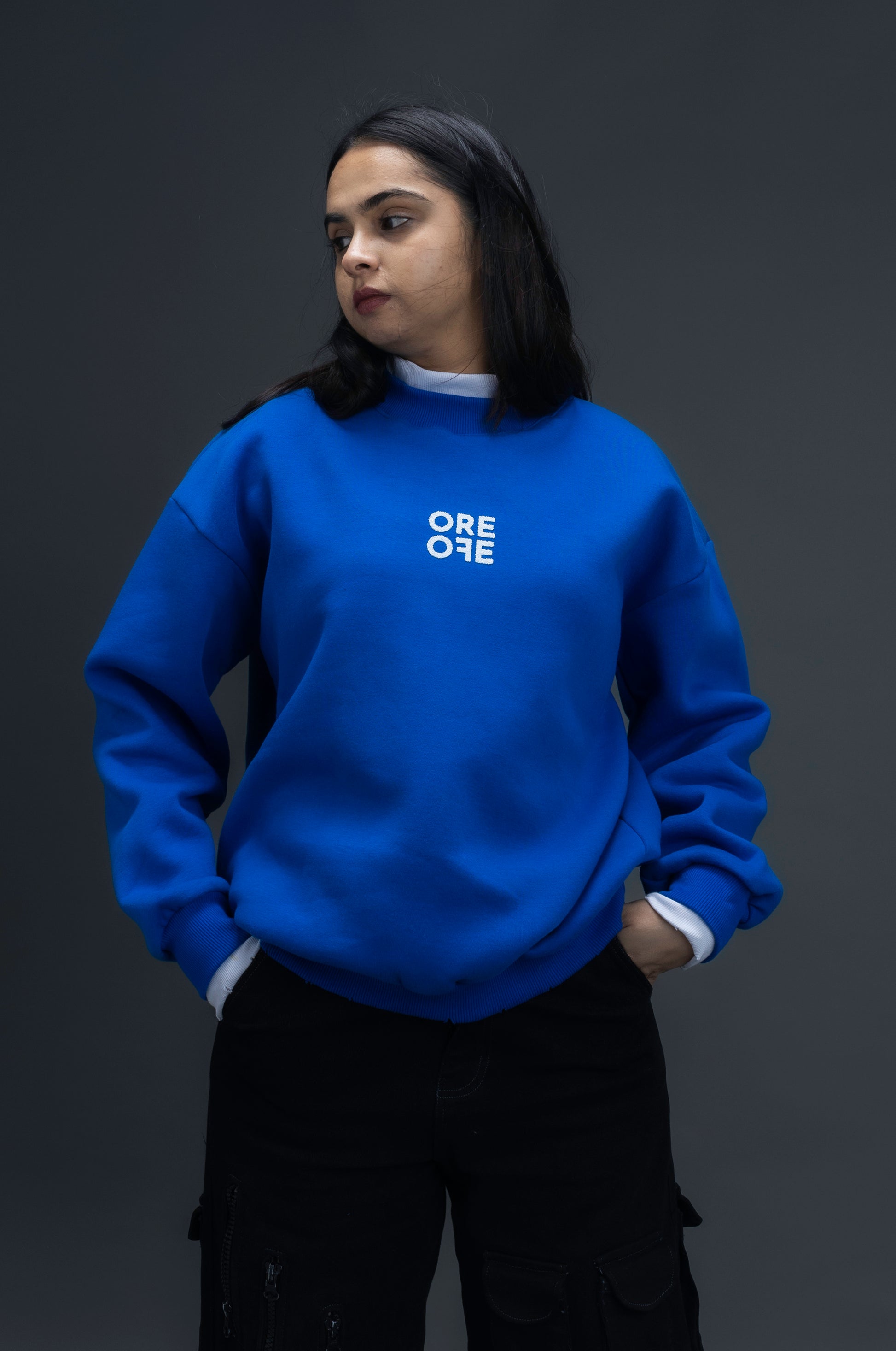 Person wearing a blue sweatshirt with ORE OFE logo on the centre of the chest standing against a grey background