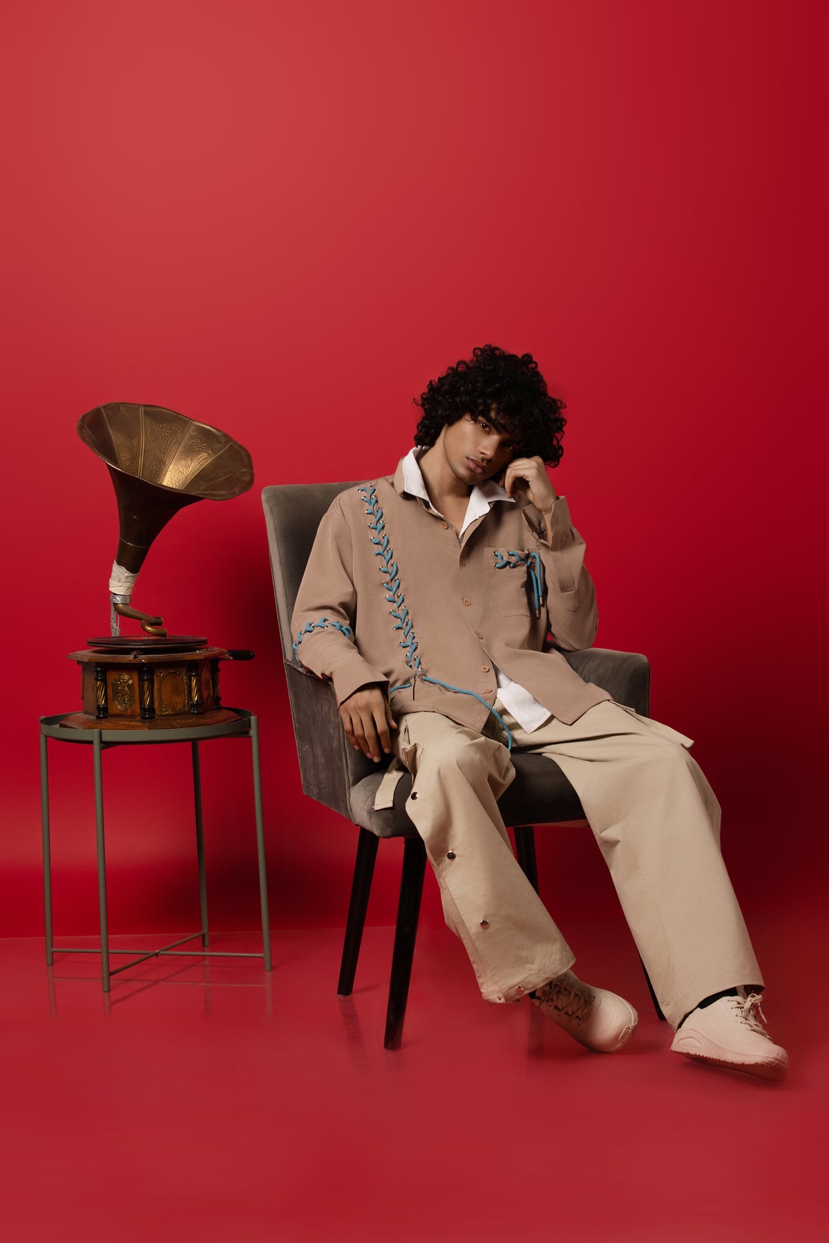 A model sitting on a chair with a vintage musical instrument next to him on a red backdrop wearing our beige and blue seoul string string and buttoned down beige pants