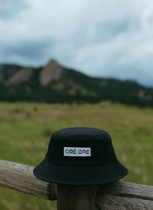 Black bucket hat with a brand logo on a wooden post against a scenic background of mountains and grasslands.