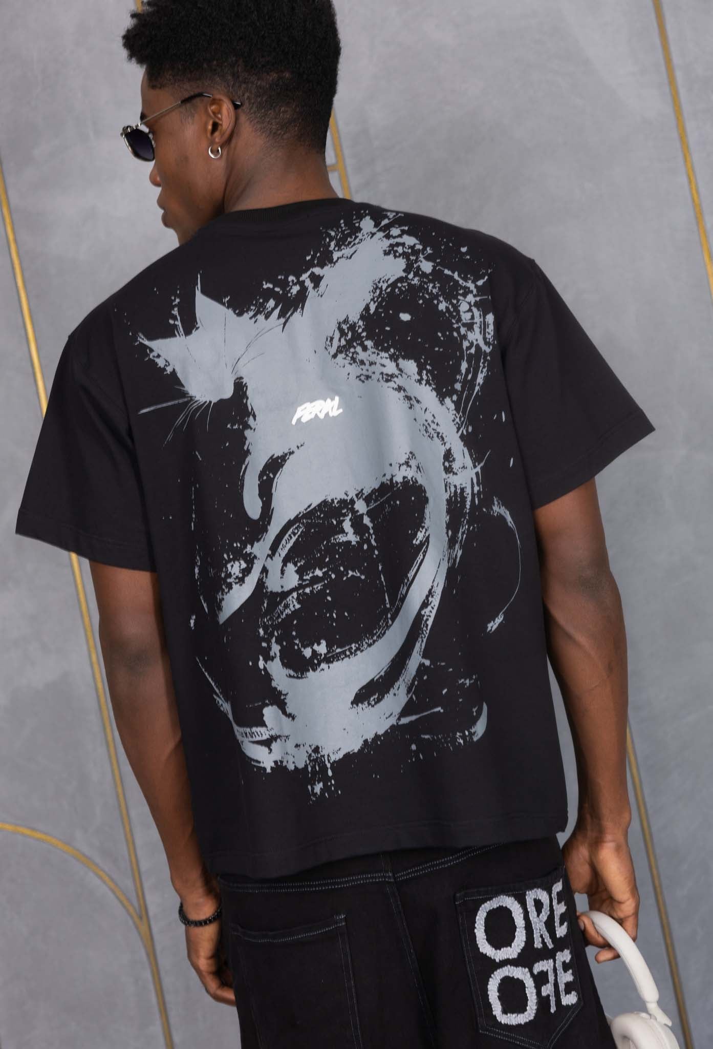 Person wearing a black t-shirt with a white graphic design on the back, standing against a gray wall.