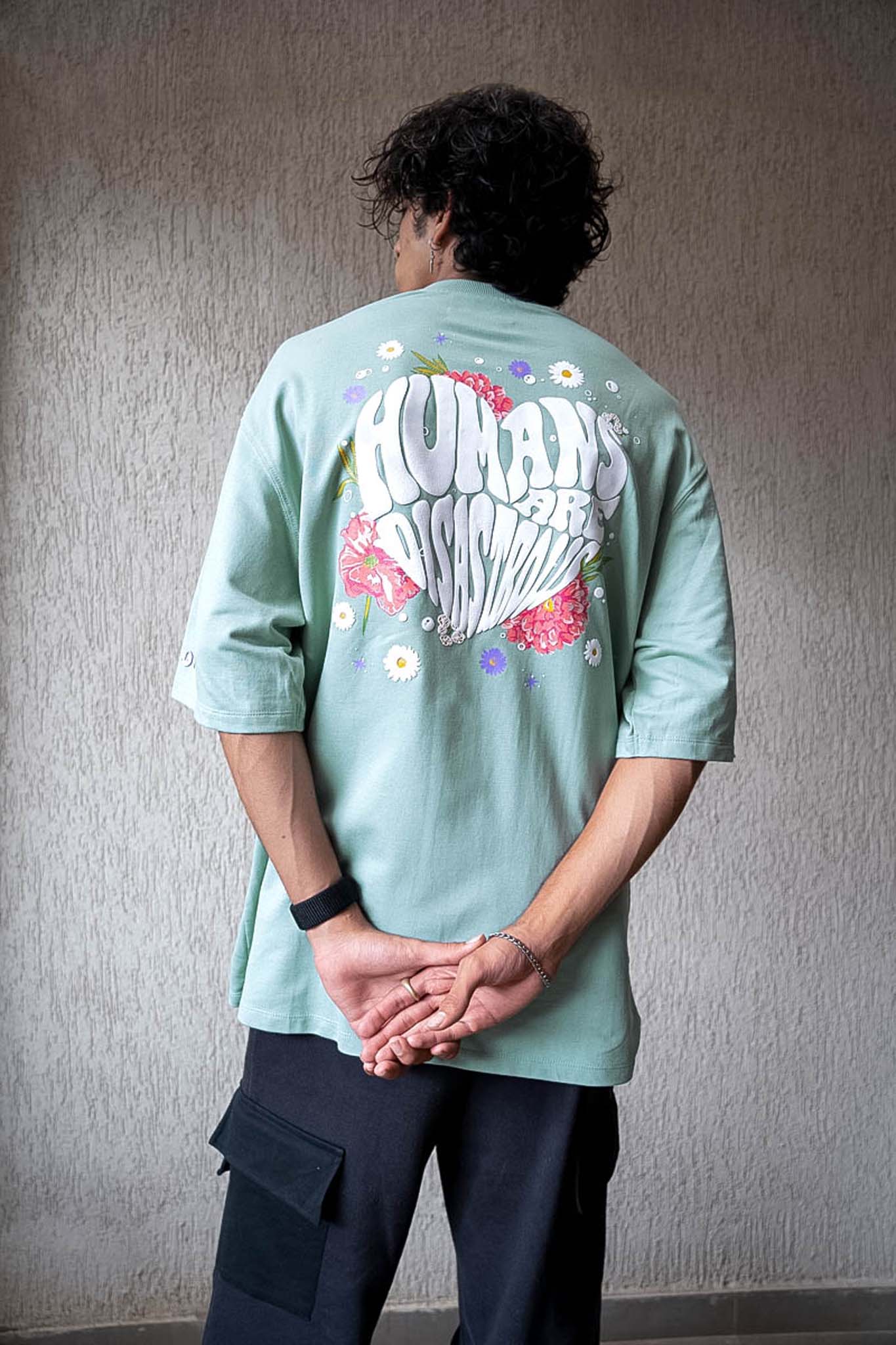 Person wearing a light green shirt with a colorful design on the back against a plain wall.