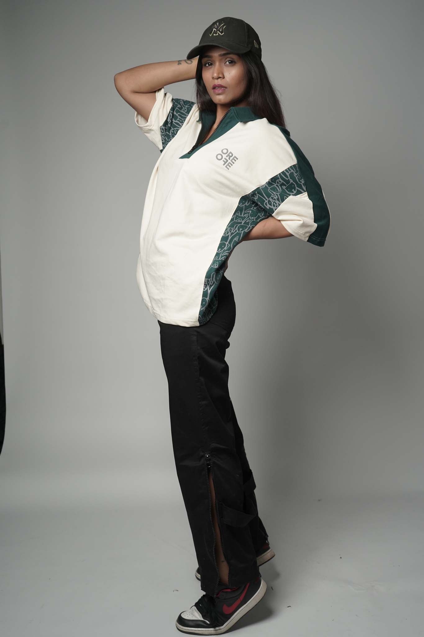 Person wearing a beige and green polo t-shirt with a logo, black pants, and a black cap on a gray background