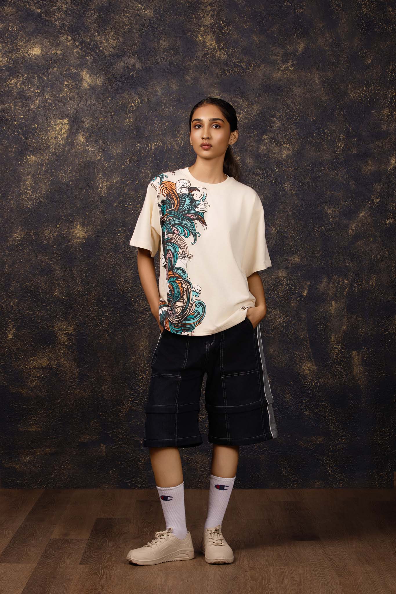 Person wearing a beige t-shirt with a colorful design and dark shorts against a textured dark background