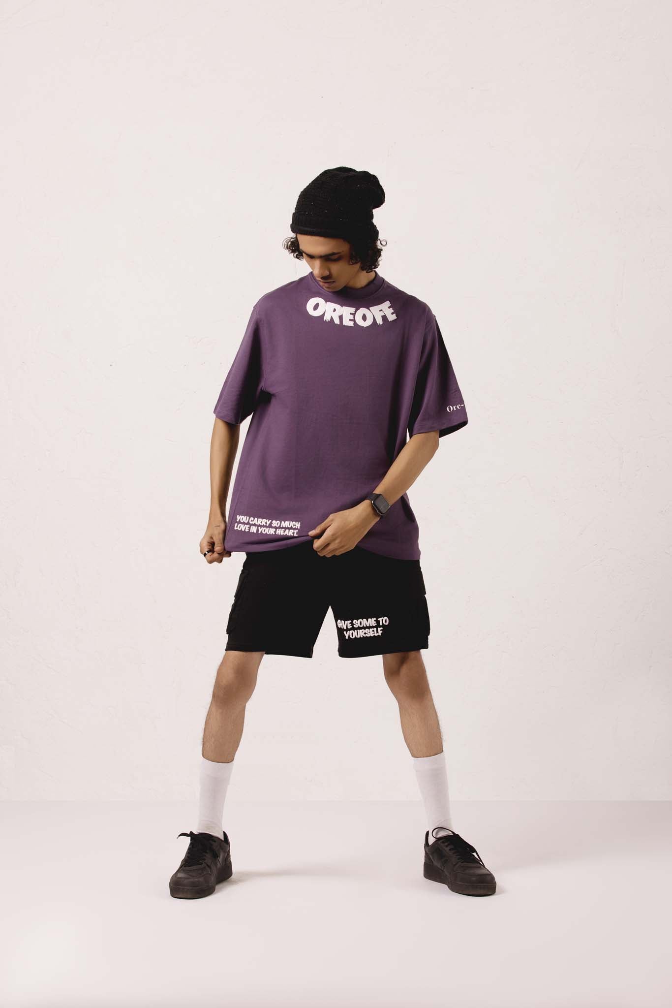 Person wearing a purple oversized t-shirt and black shorts with visible branding on a white background
