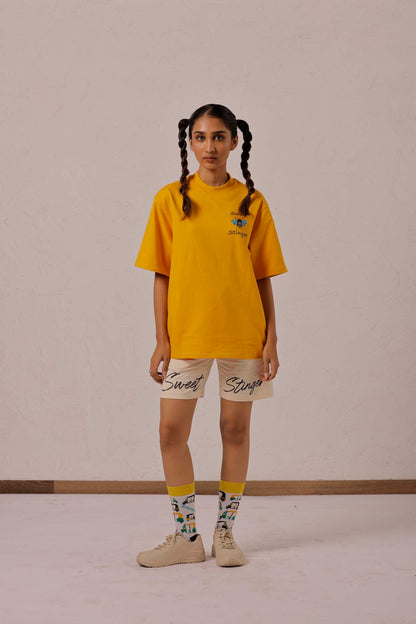 Person wearing a yellow oversized t-shirt and beige shorts with text, standing against a plain background.