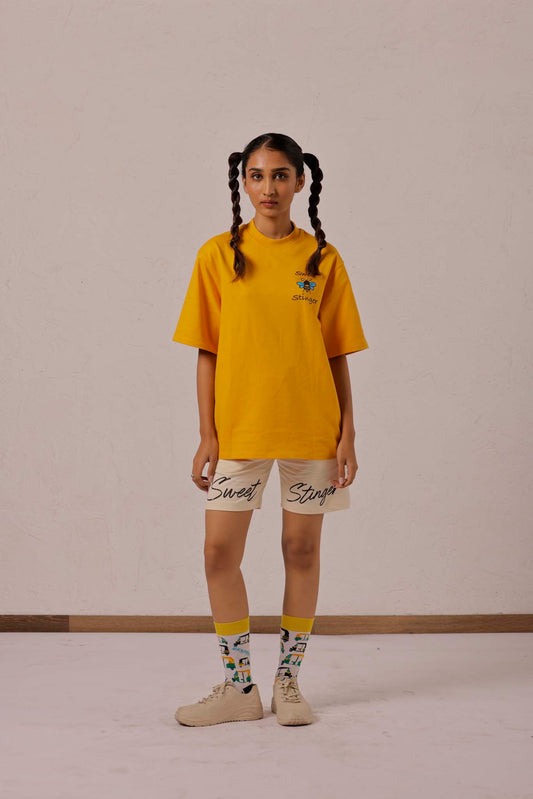 Person wearing a yellow oversized t-shirt and beige shorts with text, standing against a plain background.