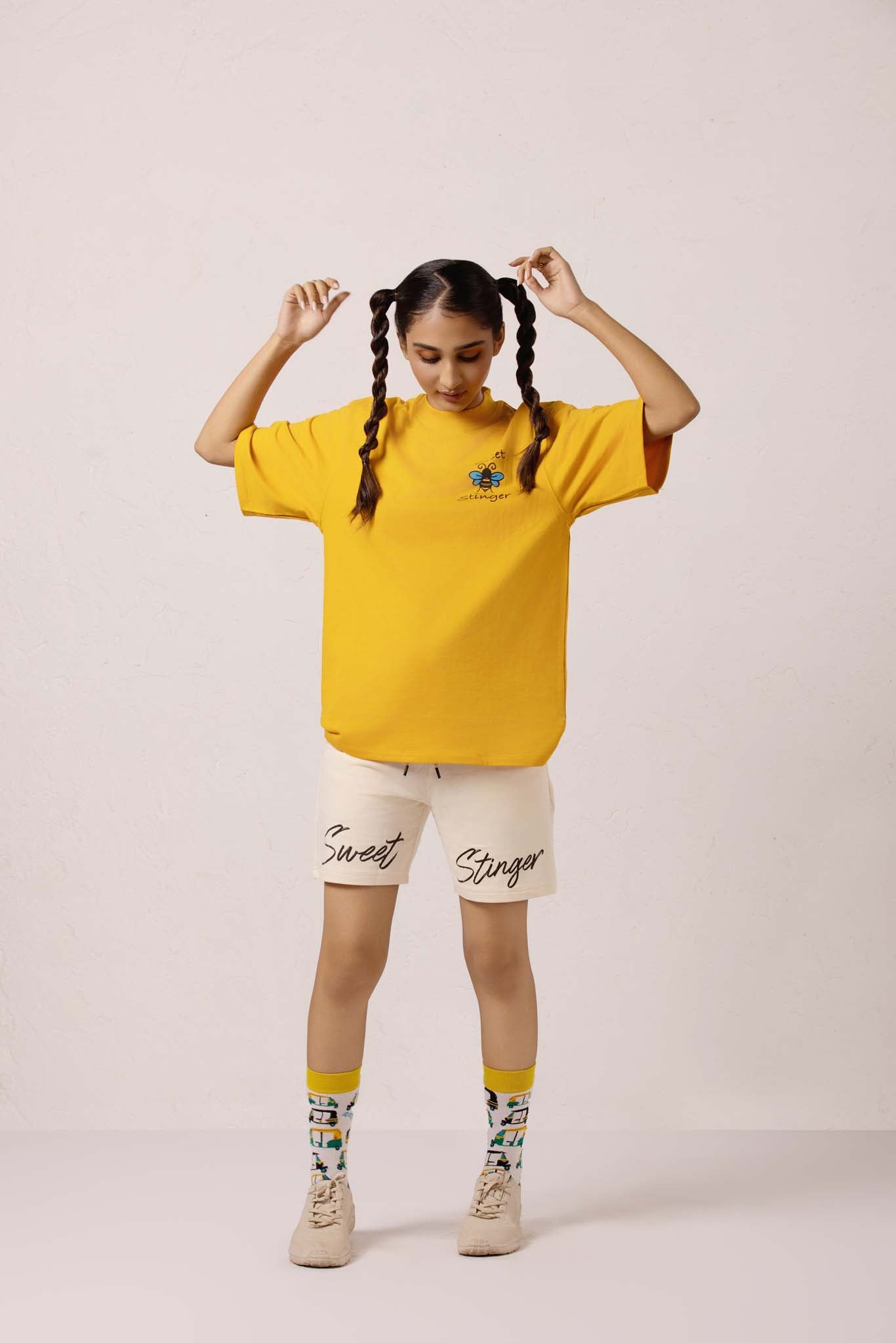 Person wearing a yellow oversized t-shirt with text, beige shorts, and colorful socks on a light pink background