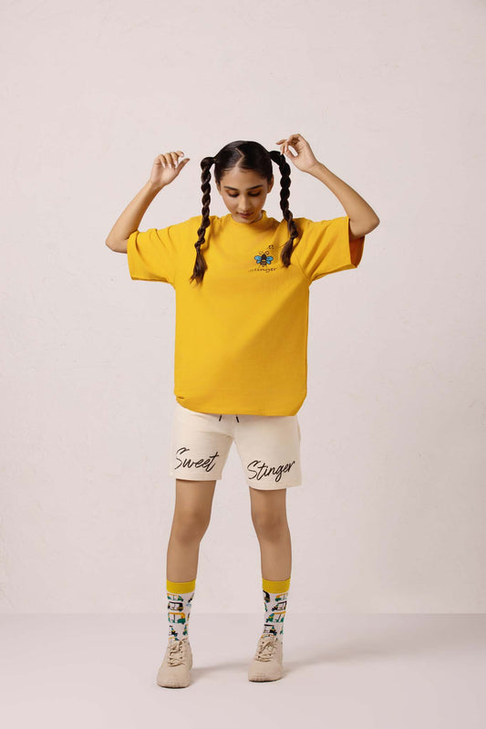 Person wearing a yellow oversized t-shirt with text, beige shorts, and colorful socks on a light pink background