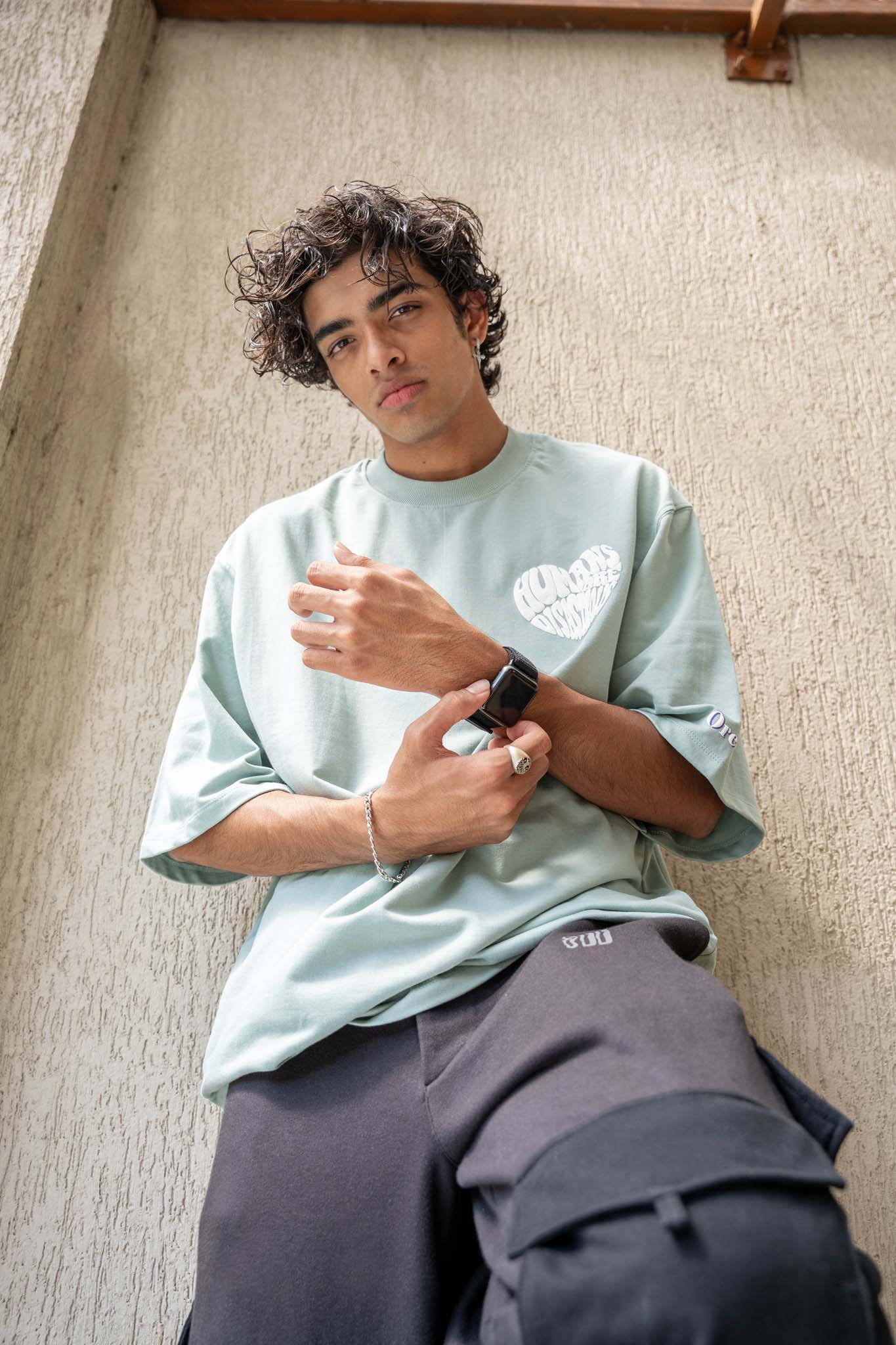 Person wearing a light green t-shirt and dark pants against a textured wall.