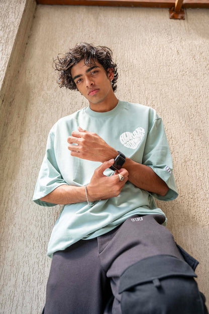 Person wearing a light green t-shirt and dark pants against a textured wall.