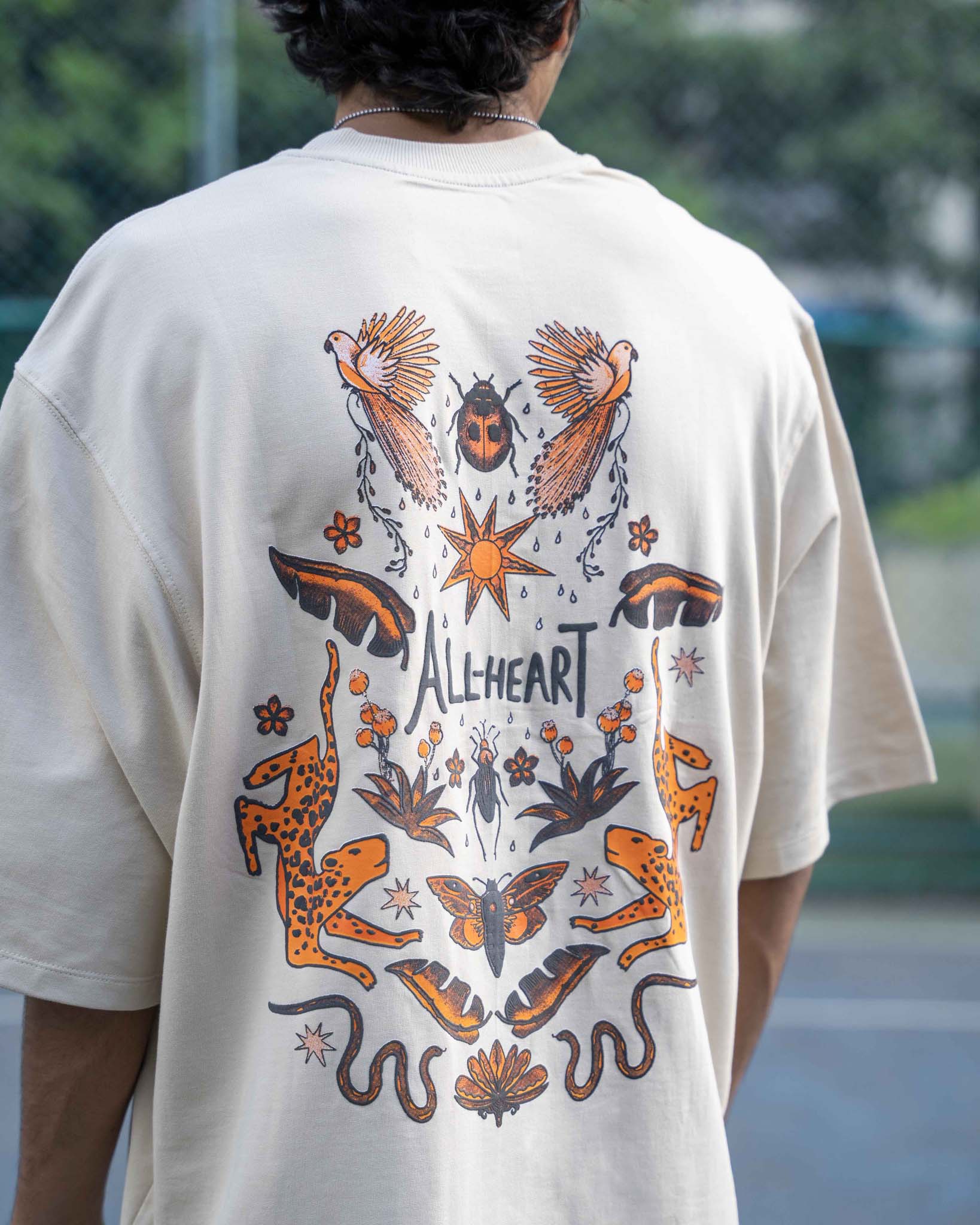 Person wearing a beige t-shirt with a colorful graphic design on the back, featuring animals and the brand 'All heart'.