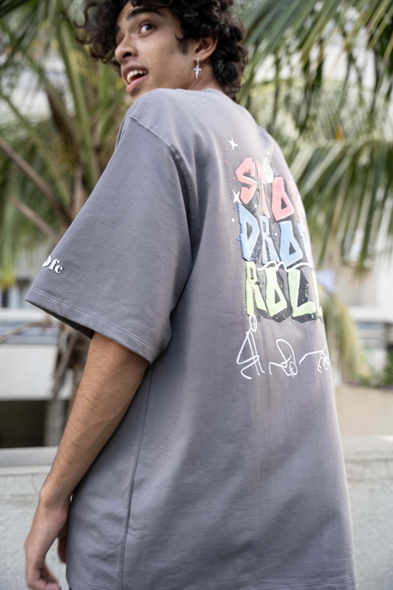 Person wearing a gray t-shirt with colorful text on the back, standing outdoors with greenery in the background.