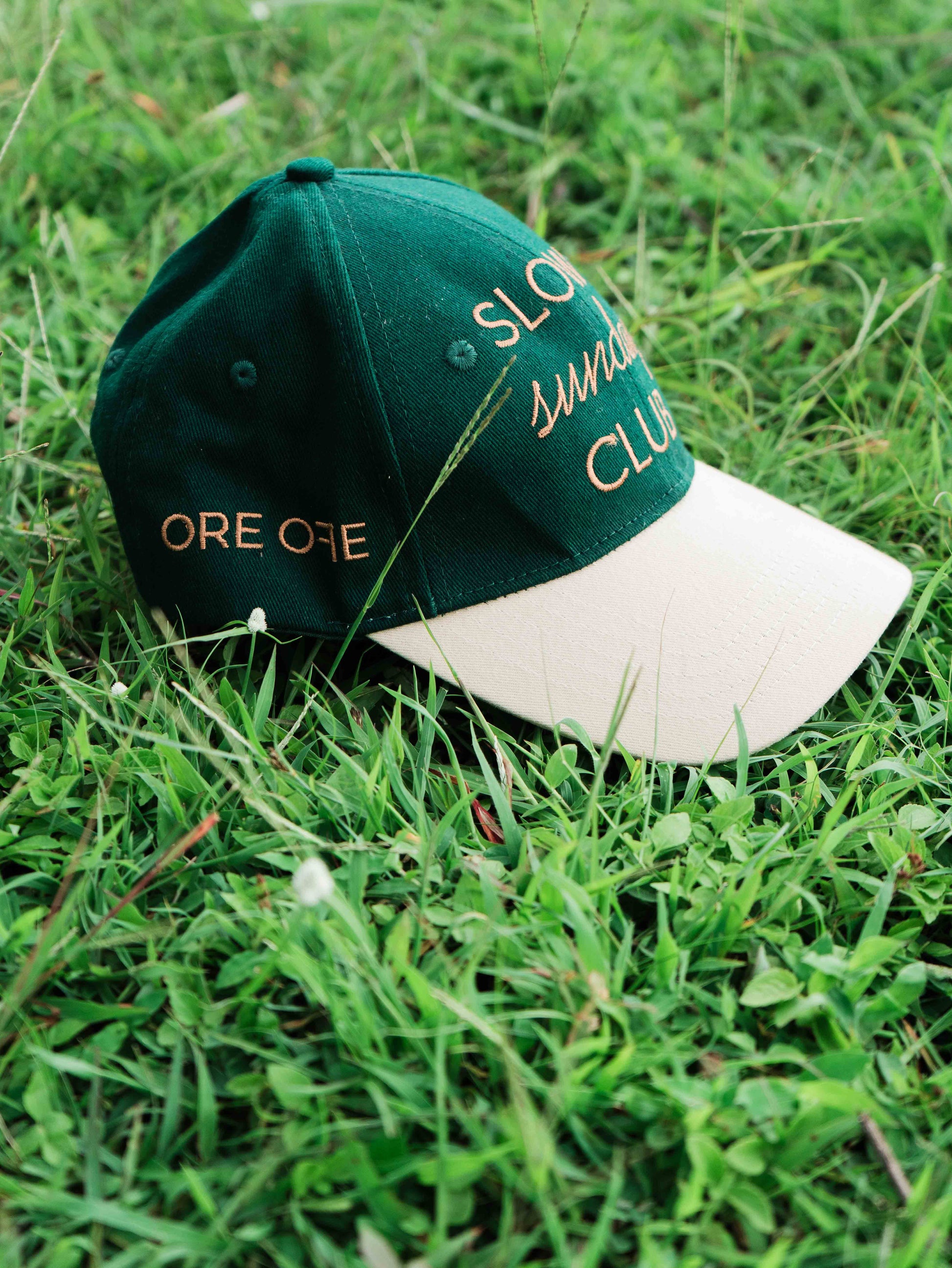 Green and white ORE OFEbaseball cap with 'Slow Sunday Club' text on grass