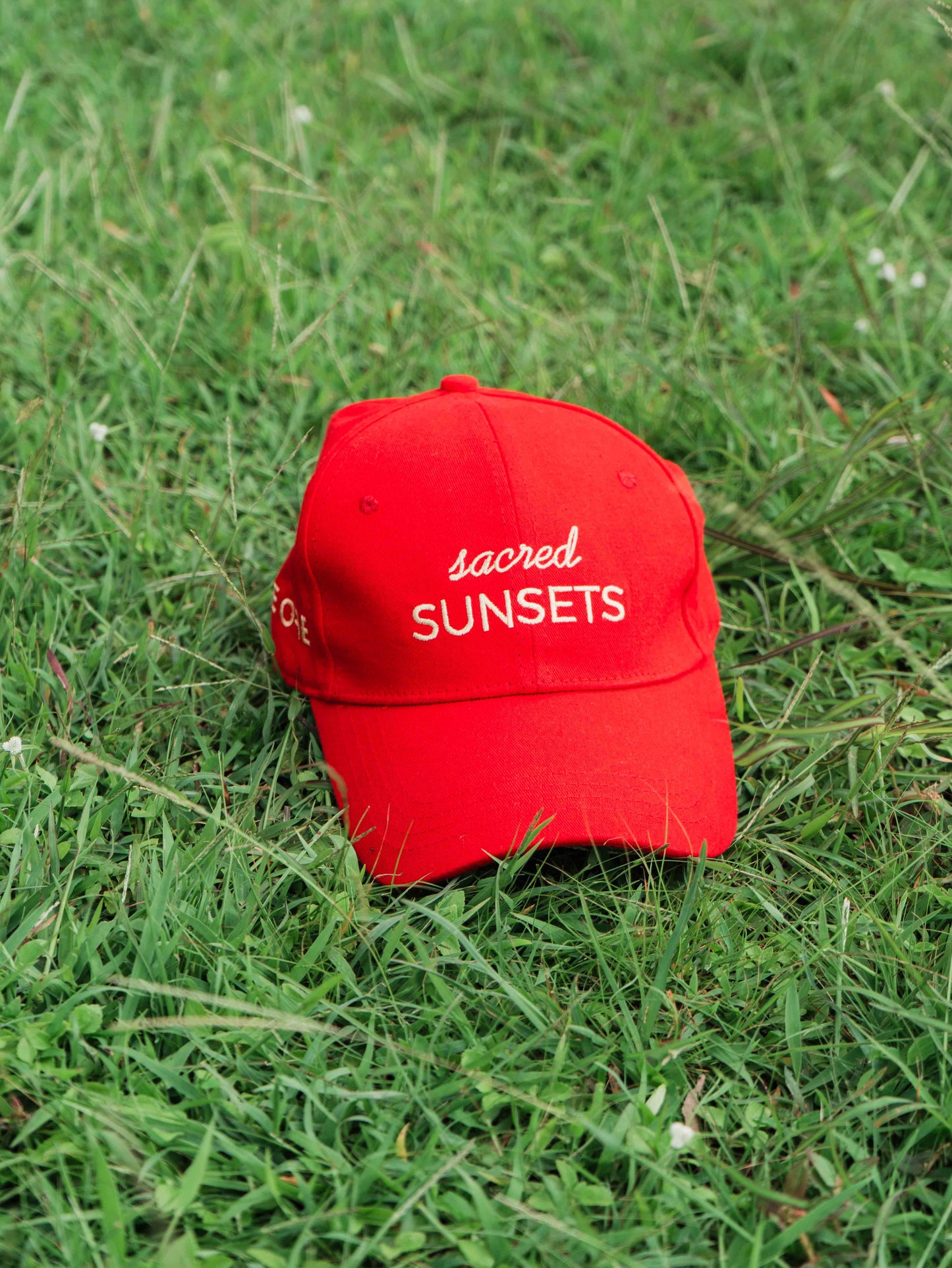 Red ORE OFE baseball cap with 'sacred SUNSETS' text on grass