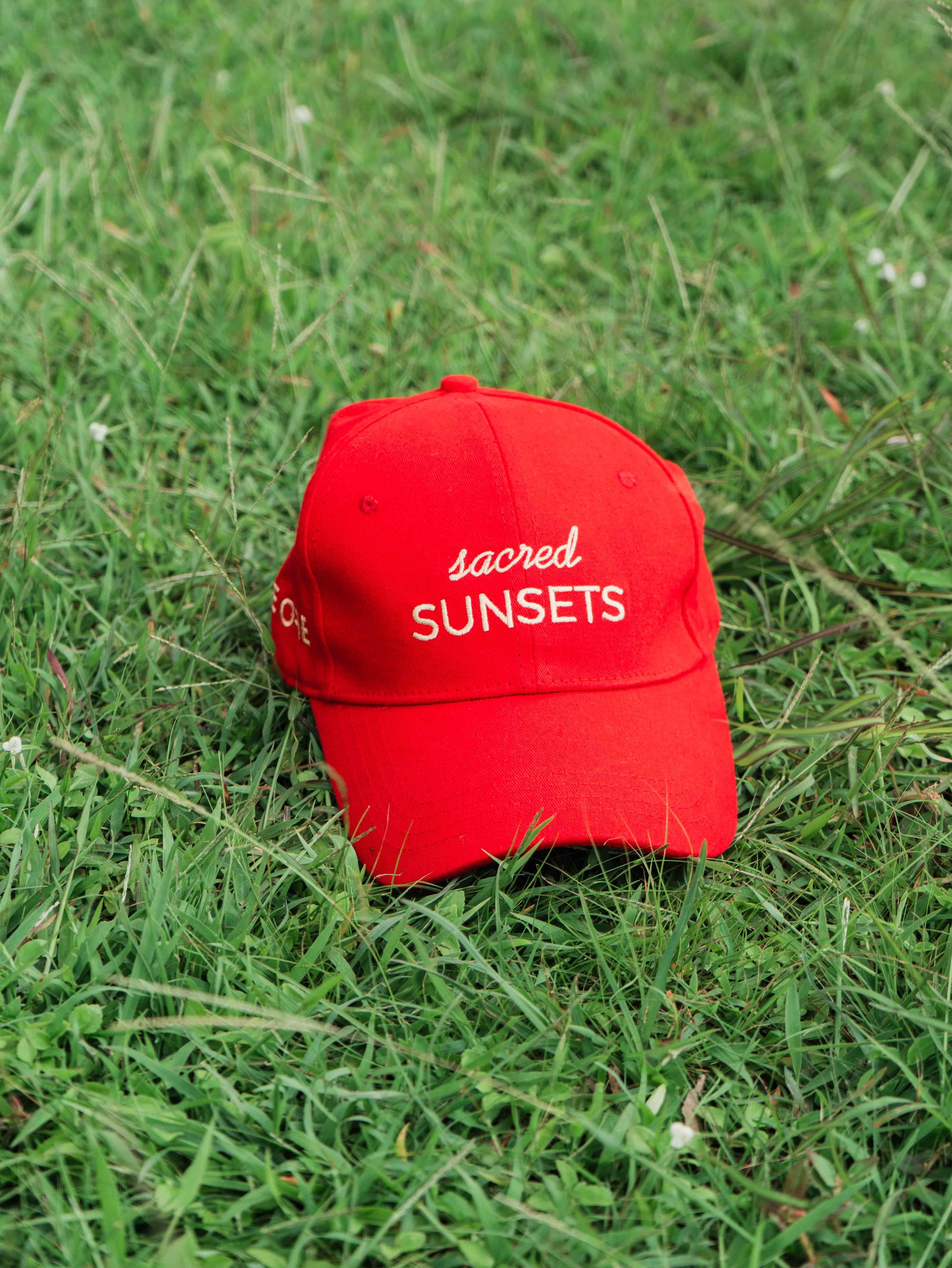 Red ORE OFE baseball cap with 'sacred SUNSETS' text on grass