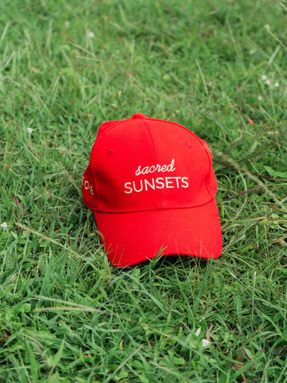 Red ORE OFE baseball cap with 'sacred SUNSETS' text on grass