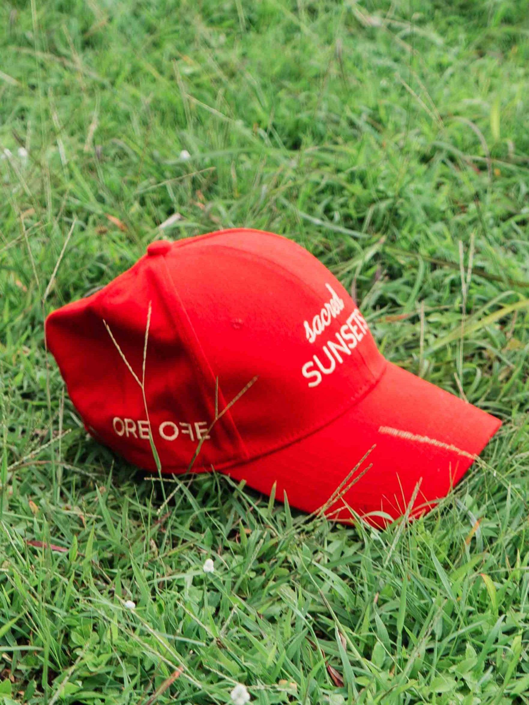 Red ORE OFE baseball cap with 'sacred SUNSETS' text on grass