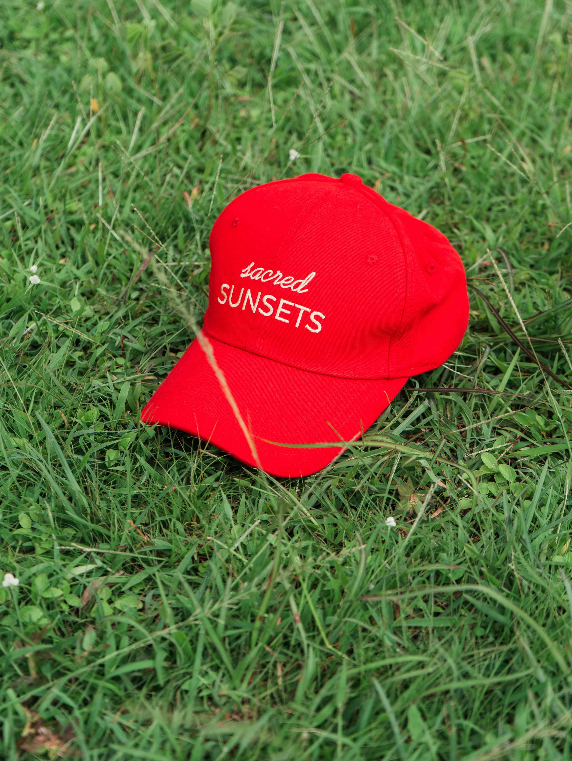 Red ORE OFE baseball cap with 'sacred SUNSETS' text on grass