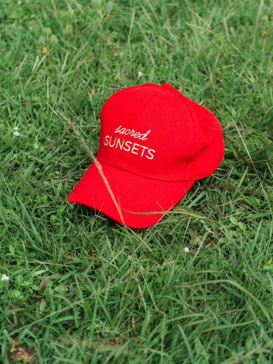 Red ORE OFE baseball cap with 'sacred SUNSETS' text on grass